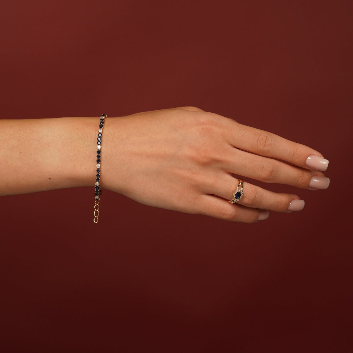 A hand wearing a delicate gold chain bracelet with small dark blue stones and a matching gold ring with a blue teardrop-shaped gemstone, set against a solid dark red background.