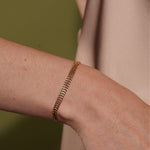 A delicate gold chain bracelet worn on a person's wrist, shown in a close-up shot against a soft olive green and beige background.