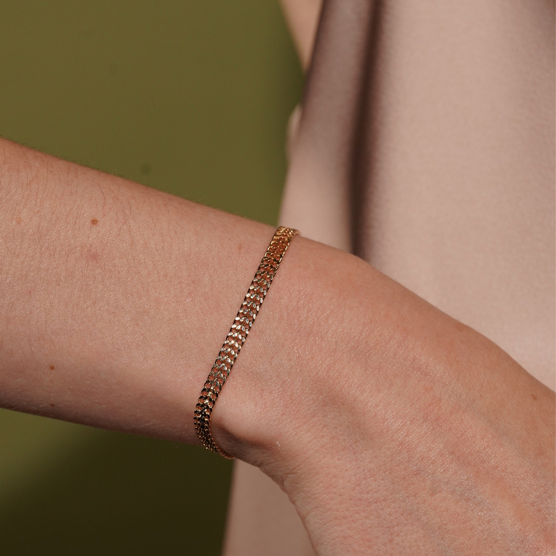 A delicate gold chain bracelet worn on a person's wrist, shown in a close-up shot against a soft olive green and beige background.