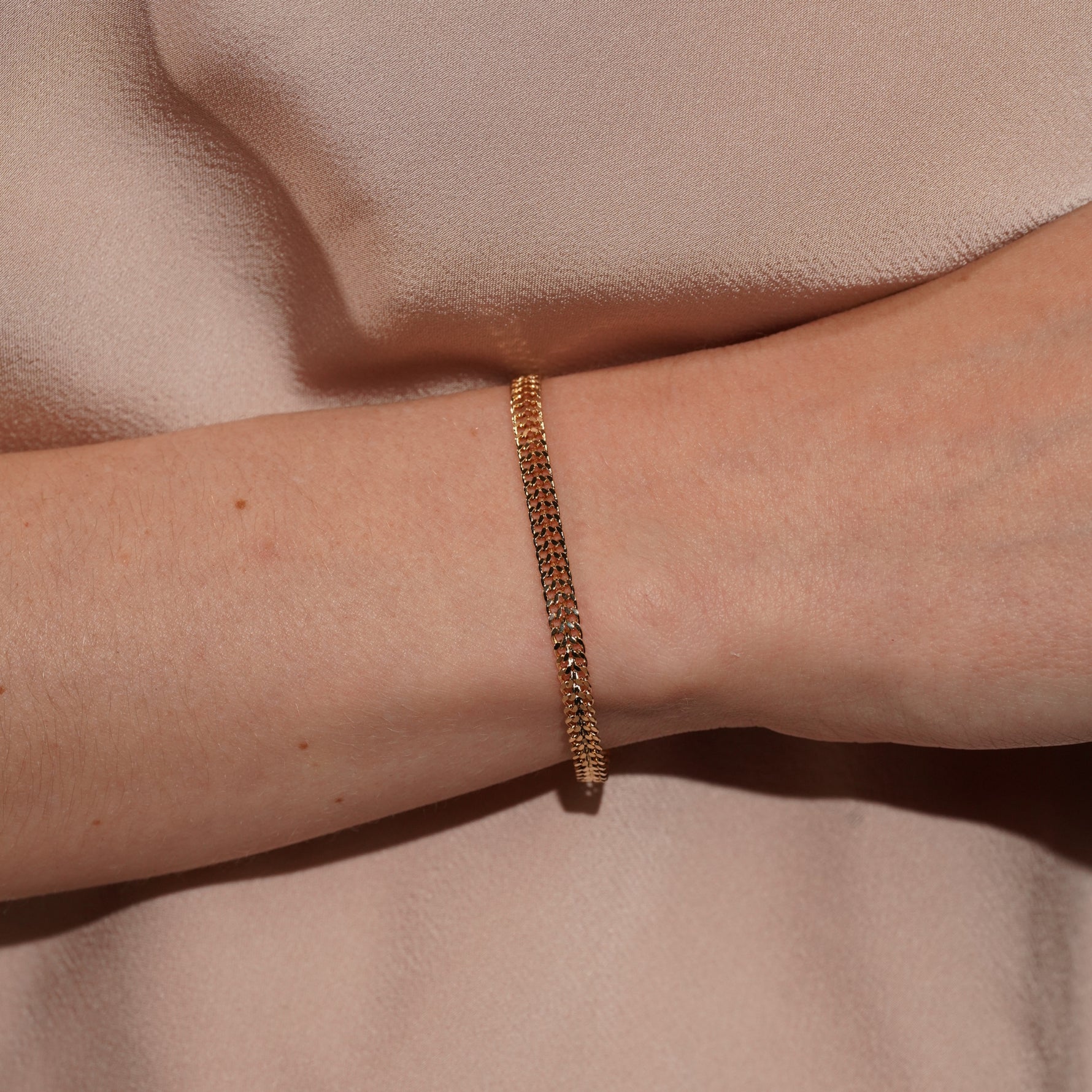 A close-up of a person's wrist wearing a delicate gold chain bracelet against a soft beige fabric background.