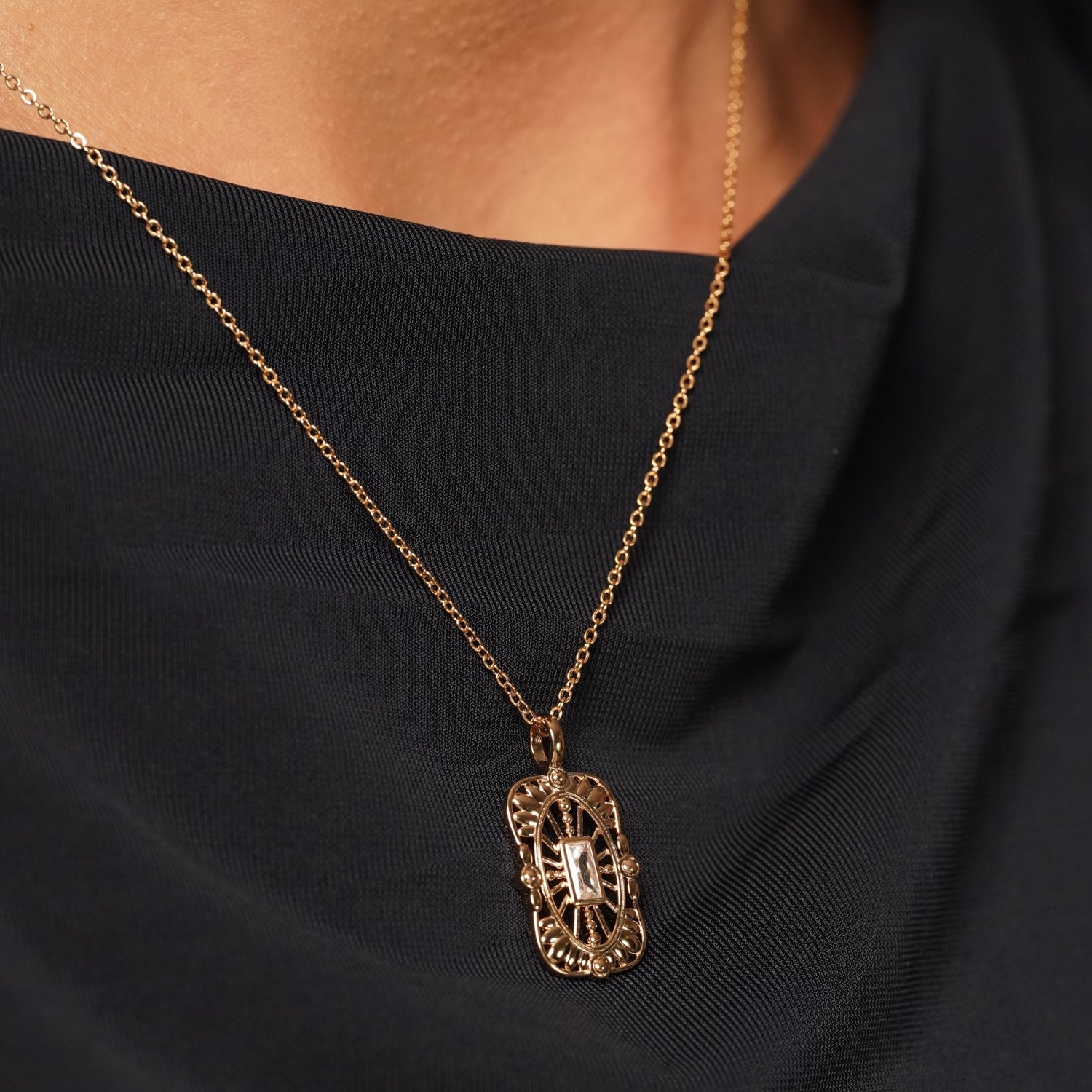 A close-up of a person wearing a delicate gold chain necklace with an ornate filigree pendant, styled against a black top.