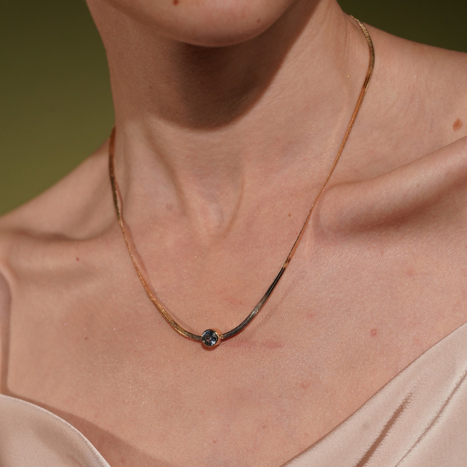 A close-up of a model's neck wearing the Marina Necklace in 18K Gold Plating Featuring a Bezel-Set Round Blue Cubic Zirconia Pendant on a Herringbone Snake Chain | Burren Jewellery