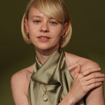 A close-up portrait of a blonde woman wearing a gold pendant necklace with a pearl-like bead and a matching gold ring, styled with a draped olive green satin scarf against a dark olive background.