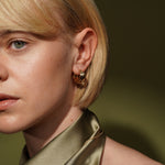 Close-up of a woman with short blonde hair wearing a gold triple-hoop earring, paired with a satin olive green collar against a dark olive background.