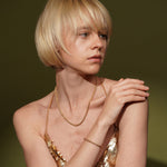 A blonde model with a short bob haircut wearing a plated gold chain necklace and a gold bracelet, posing in a metallic gold sequined top against a dark olive green background.