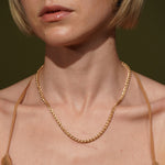 A close-up of a model wearing a delicate plated gold chain necklace with a textured, woven or braided appearance. The shot focuses on the necklace against the model's neck and upper chest, with a solid dark olive green background.