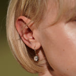A close-up side profile of a person's ear wearing a small gold drop earring with a round bezel-set clear stone.