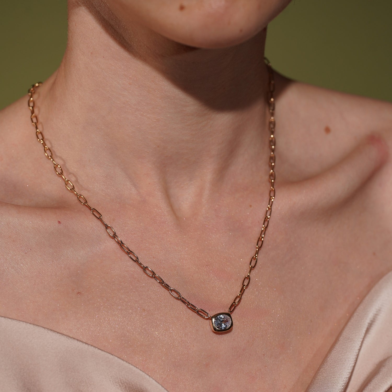 A close-up of a model's neck and upper chest wearing a delicate plated gold chain necklace with a small bezel-set oval gemstone pendant.