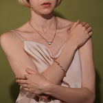 A close-up lifestyle shot of a model wearing a delicate plated gold chain necklace with a small clear stone pendant, a matching gold bracelet, and a gold ring with a clear stone, styled with a pale pink satin top against an olive green background.