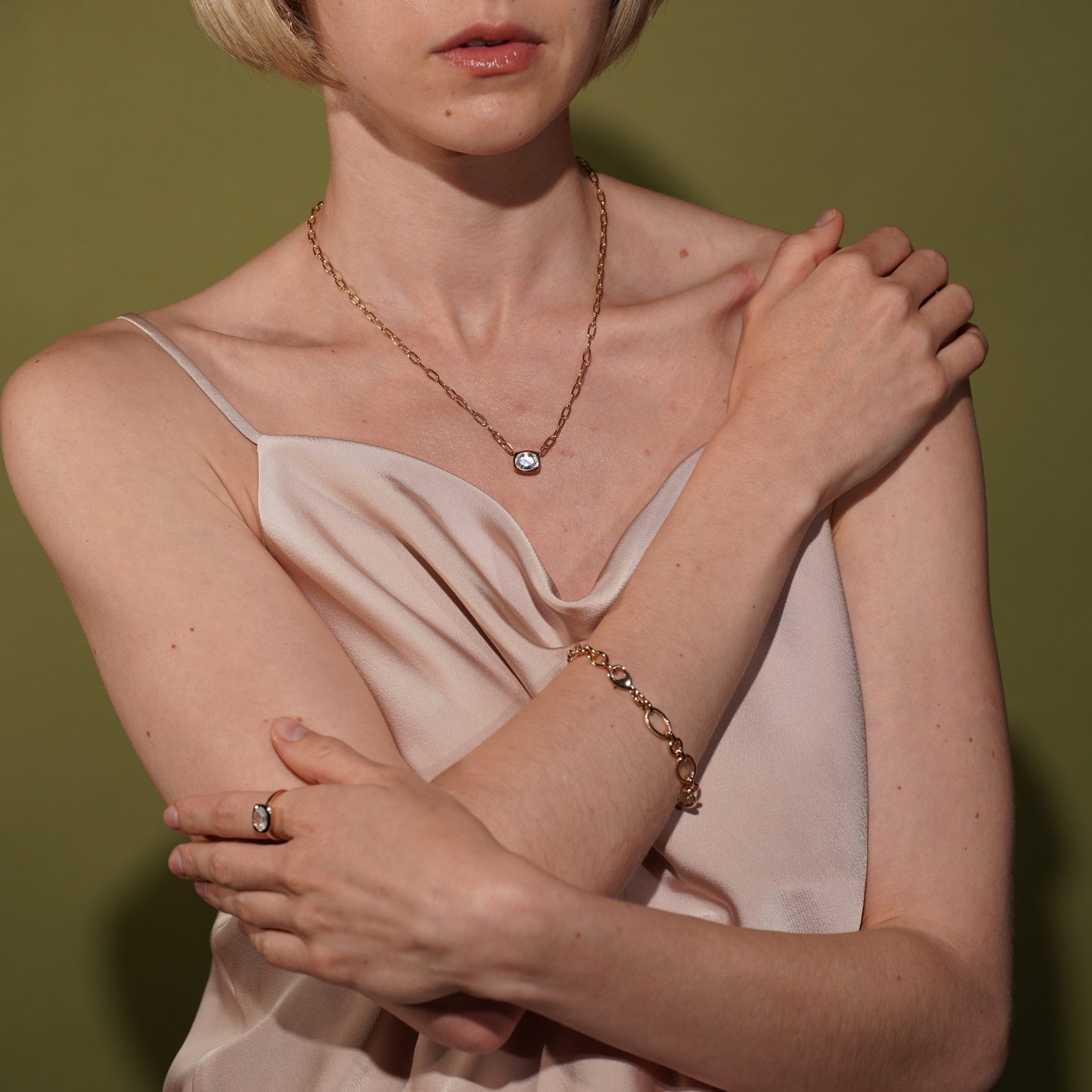 A close-up lifestyle shot of a model wearing a delicate plated gold chain necklace with a small clear stone pendant, a matching gold bracelet, and a gold ring with a clear stone, styled with a pale pink satin top against an olive green background.