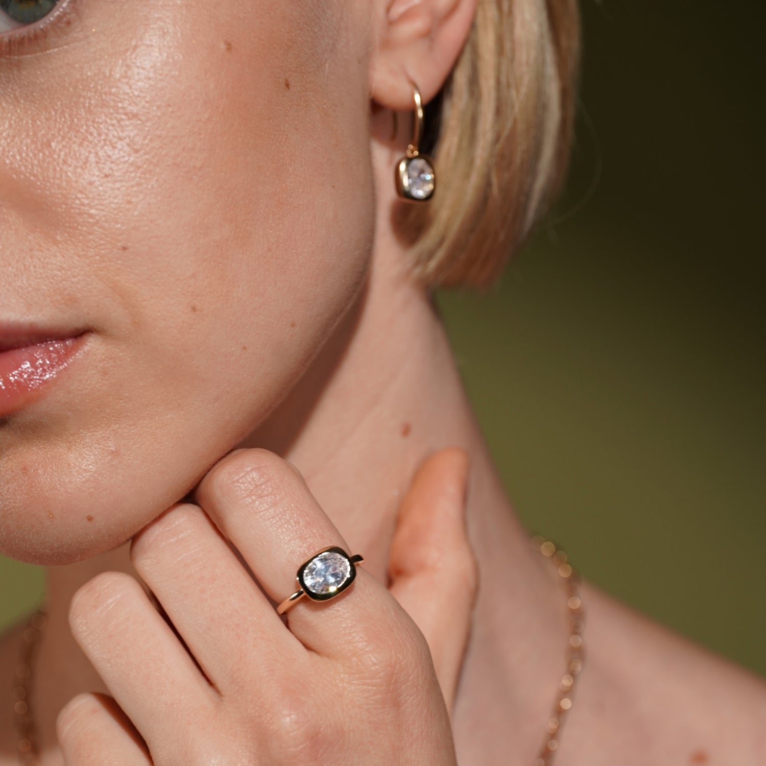 Close-up of a woman wearing a gold ring with a clear oval gemstone on her finger and a matching earring, with a blurred olive-green background.