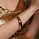 A close-up of a person's arm wearing a polished gold plated Burren Jewellery bangle bracelet, with a shimmering gold sequined top in the background.