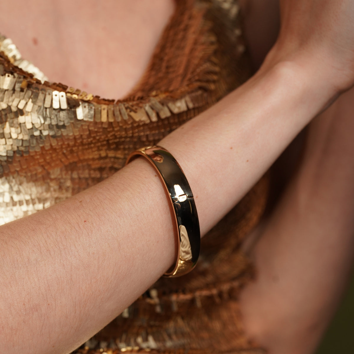 A close-up of a person's arm wearing a polished gold plated Burren Jewellery bangle bracelet, with a shimmering gold sequined top in the background.