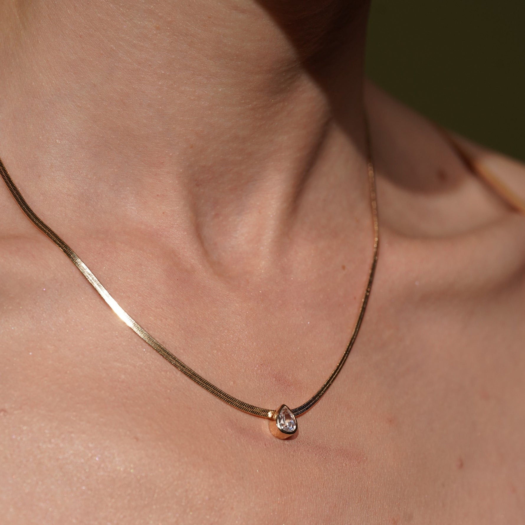 A close-up of Model's Neck wearing Date Night Necklace in 18K Gold Plating Featuring a Bezel-Set Pear Cut Clear Cubic Zirconia Pendant on a Herringbone Snake Chain | Burren Jewellery