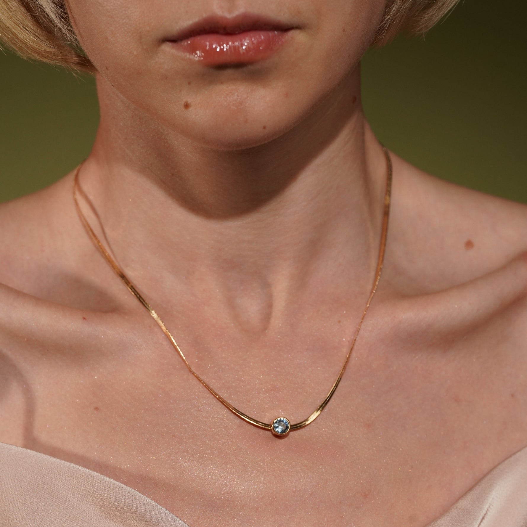 A close-up of a model's neck and upper chest wearing Marina Necklace in 18K Gold Plating Featuring a Bezel-Set Round Blue Cubic Zirconia Pendant on a Herringbone Snake Chain | Burren Jewellery