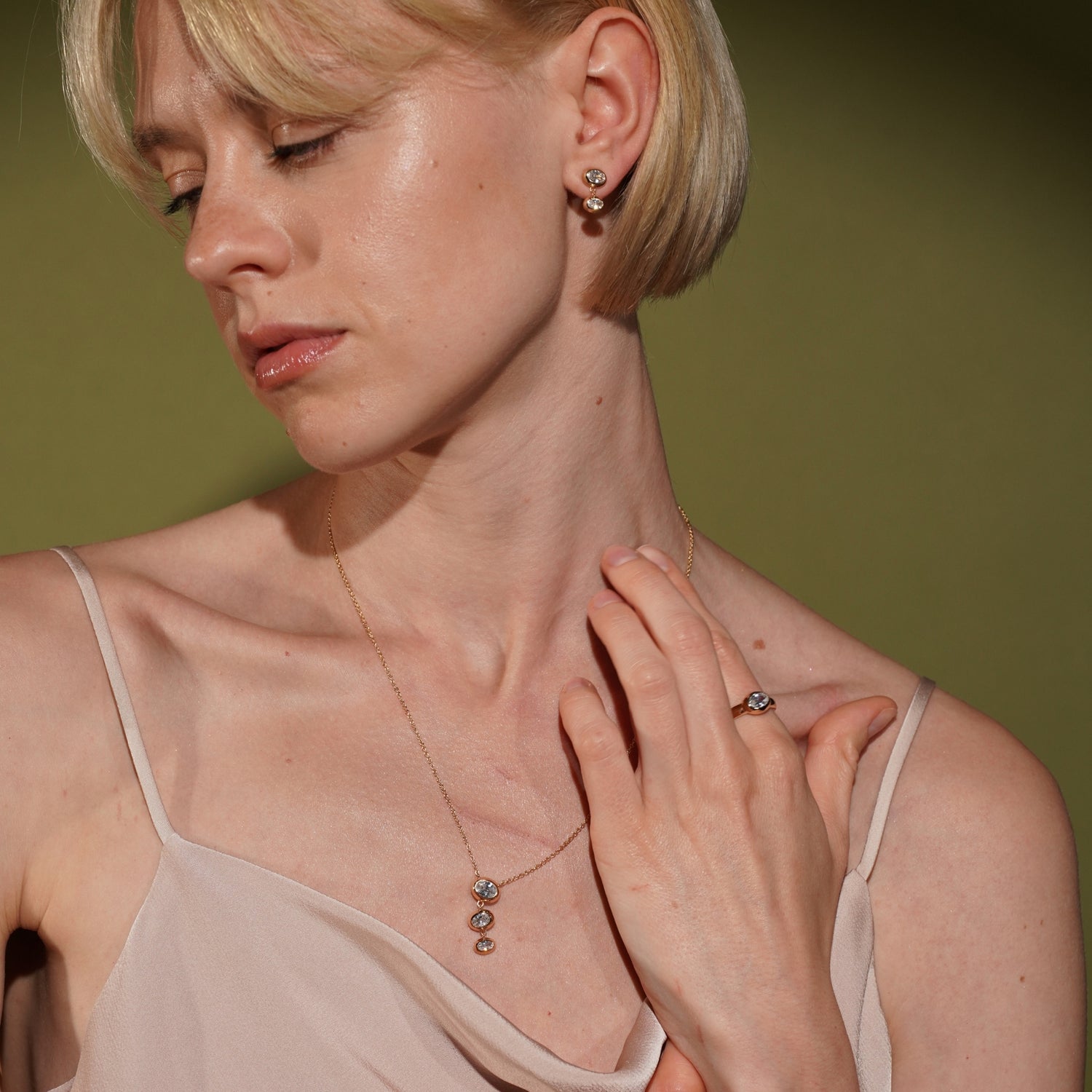 A close-up lifestyle shot of a blonde woman wearing a delicate gold necklace with matching ring and stud earrings