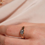 A close-up of a hand wearing a gold ring with a textured, stone-like oval center, set against a soft beige fabric background.
