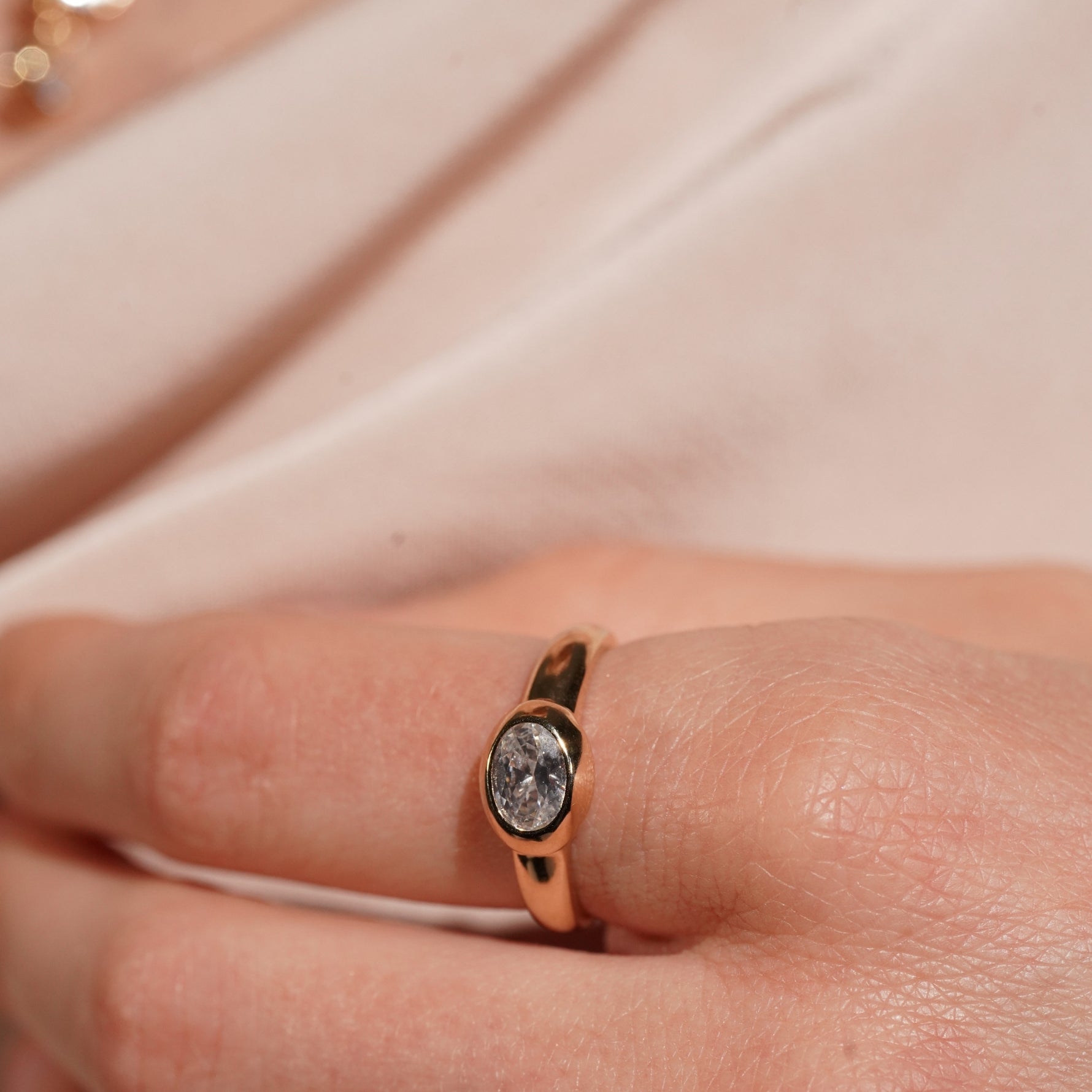 A close-up of a hand wearing a gold ring with a textured, stone-like oval center, set against a soft beige fabric background.