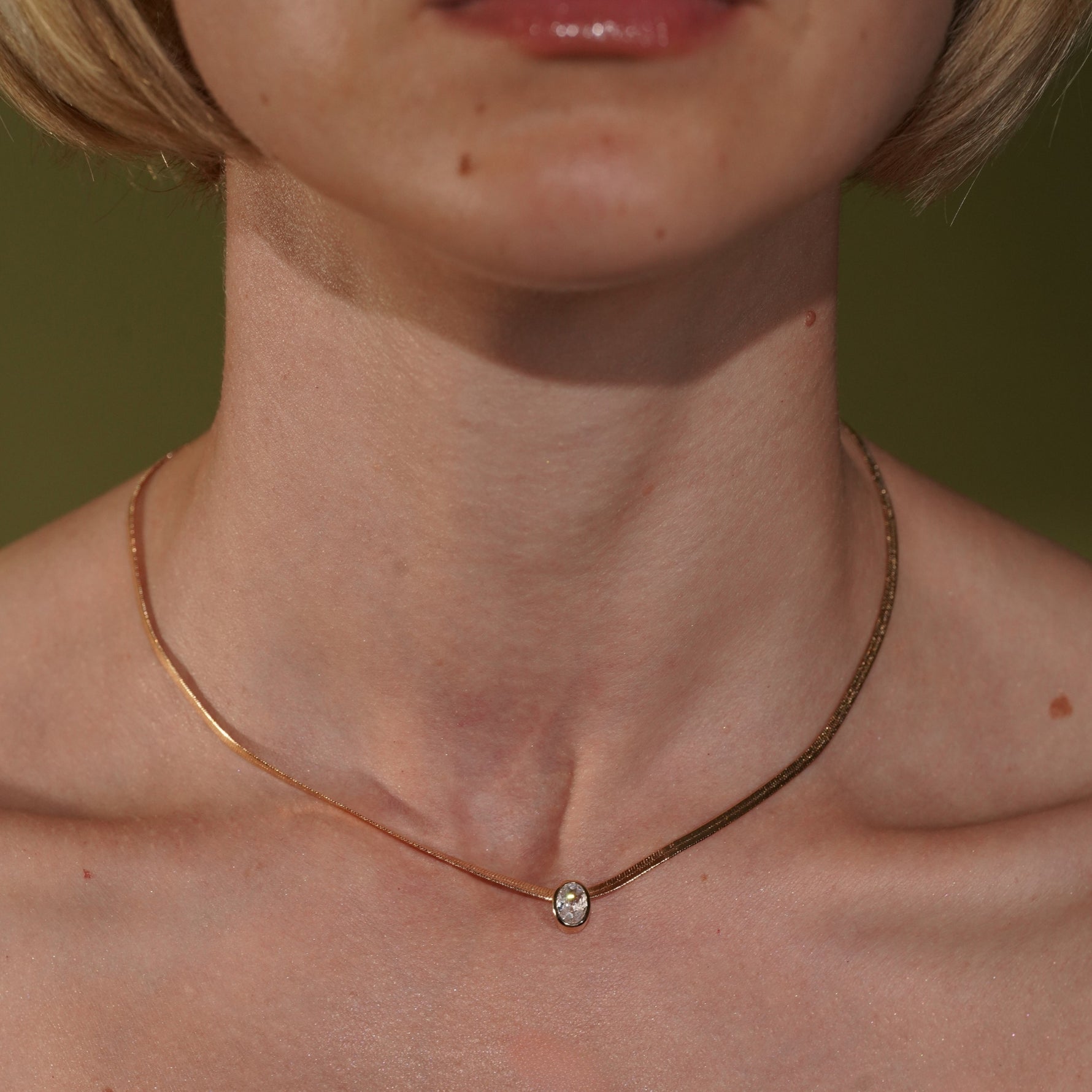 A close-up of a model's neck and collarbone wearing a delicate gold chain necklace with a single small clear gemstone pendant.