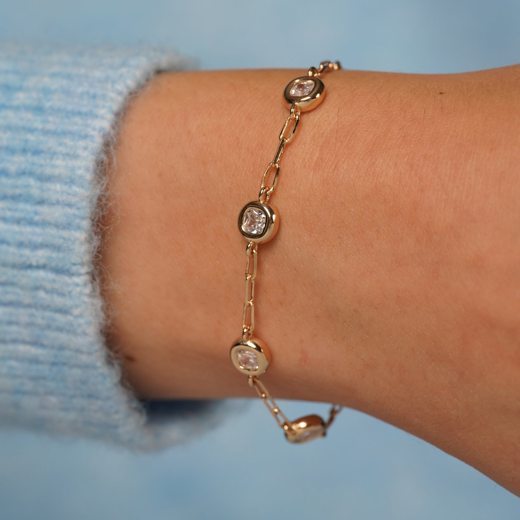 A delicate gold chain bracelet with bezel-set clear round stones, shown worn on a person's wrist with a light blue knit sleeve visible.