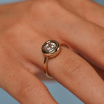 A close-up of a hand wearing a rose gold ring with a bezel-set cushion-cut clear gemstone, set against a soft blue background.