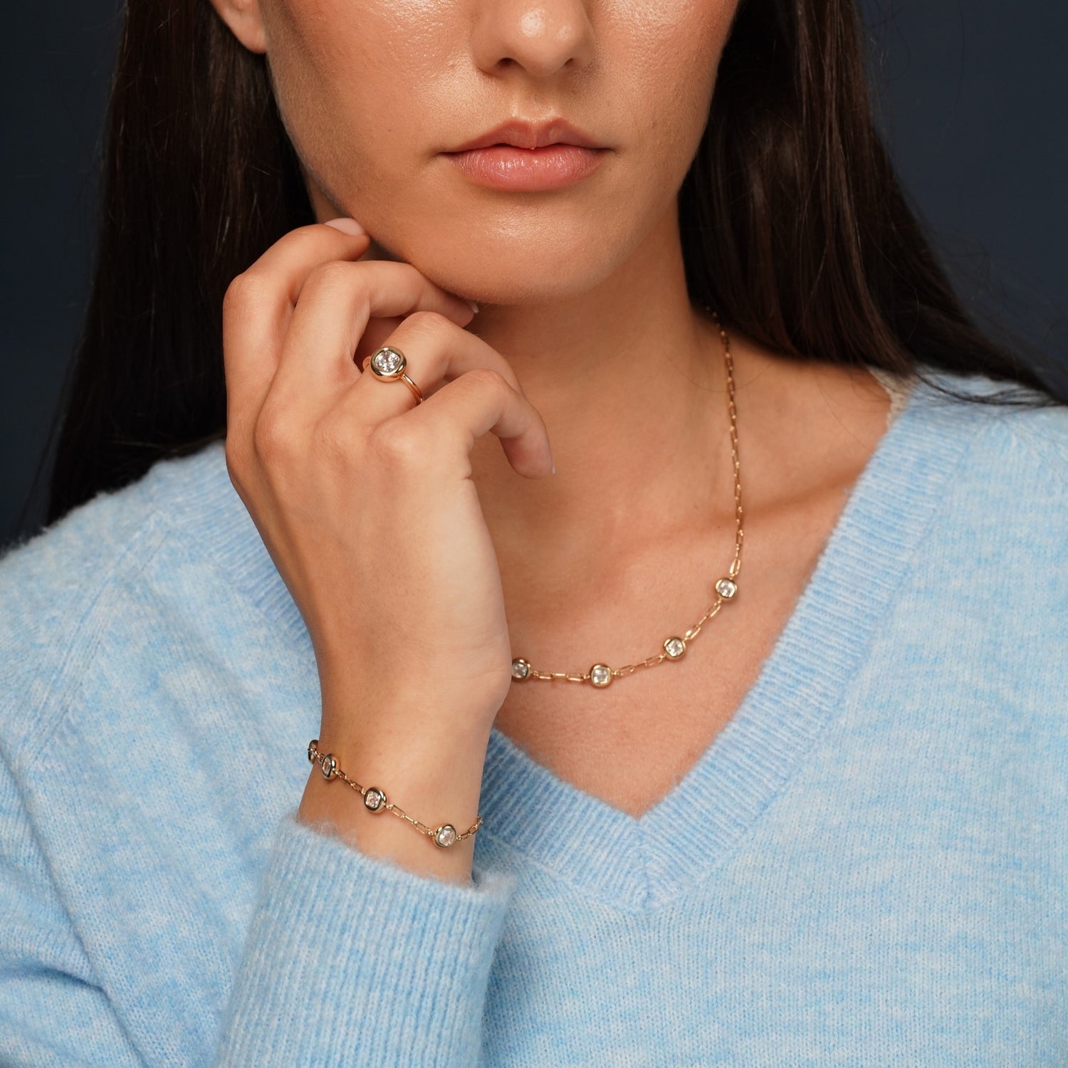 A close-up lifestyle shot of a model wearing a delicate gold necklace and matching bracelet and ring, each adorned with small clear crystals.