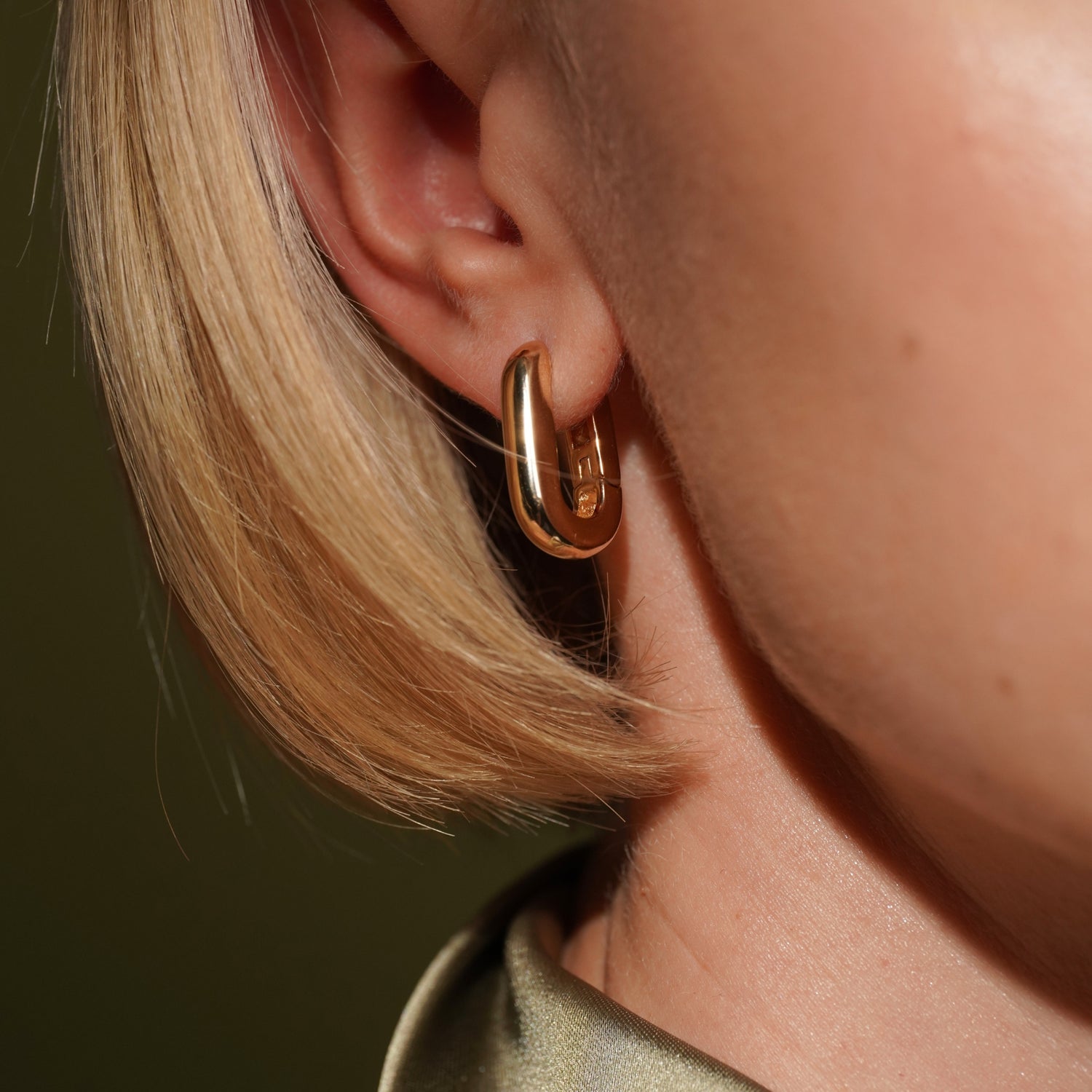 A close-up of a person's ear wearing a polished champagne gold colour huggie hoop earring, with blonde hair and skin visible against a dark olive background.