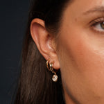 A close-up of a model wearing Timeless Solitaire Earrings in 18K Gold Plating Featuring Oval-Cut Cubic Zirconia Drop Charms on Polished Huggie Hoops | Burren Jewellery