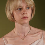 A close-up portrait of a woman with short blonde hair wearing a delicate gold chain necklace with a small clear stone pendant, set against a solid olive green background.