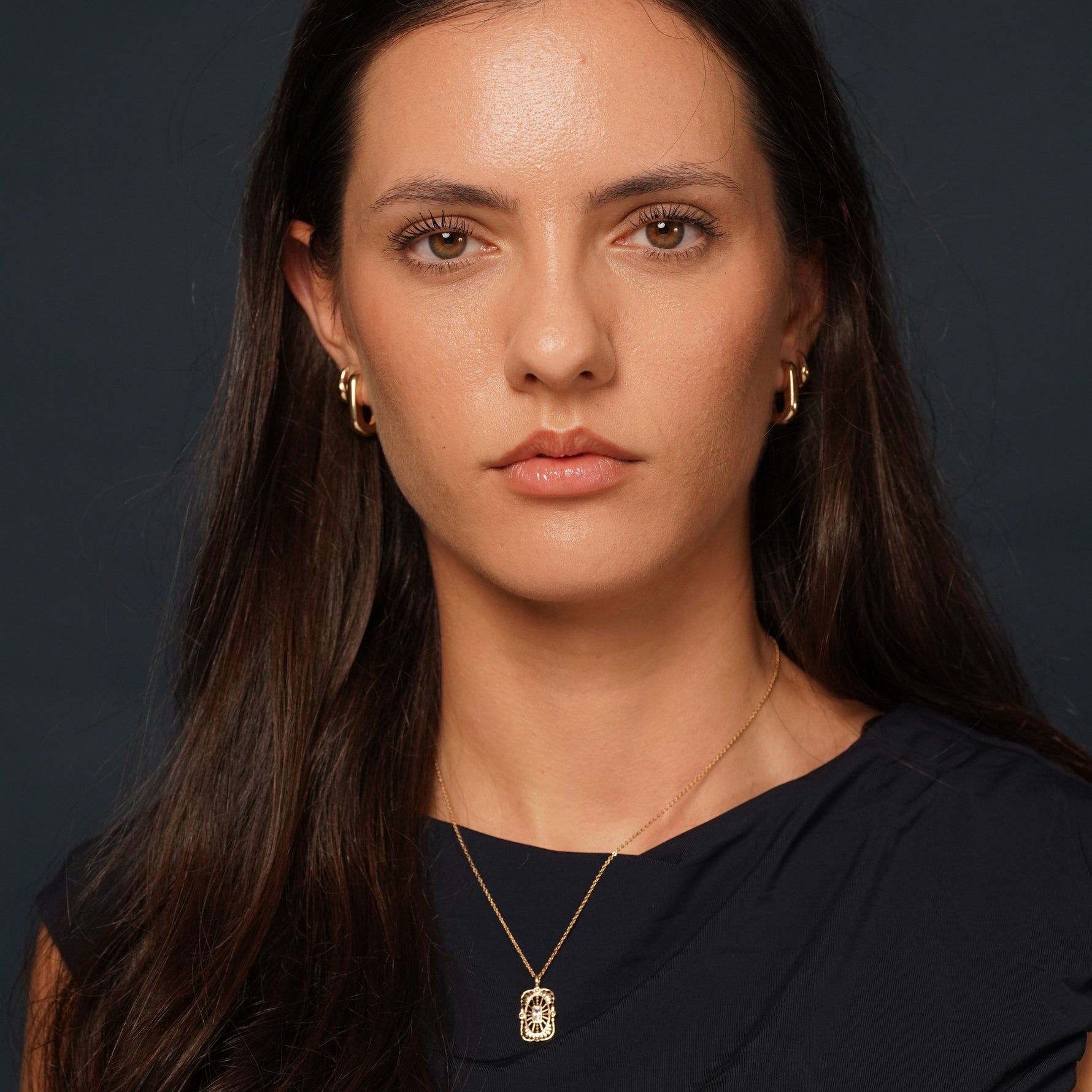 A close-up portrait of a woman with long dark hair wearing a delicate gold necklace with a rectangular pendant and matching small gold hoop earrings