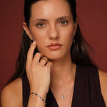 A close-up portrait of a woman wearing delicate jewelry, including a small gold ring, a delicate gold necklace with a small green and white pendant, and a beaded bracelet, set against a solid dark reddish-brown background.