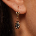 A close-up of a gold drop earring worn on a person's ear, featuring a marquise-shaped dark blue stone surrounded by a gold halo.