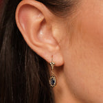 A close-up of a person's ear wearing a gold drop earring with an oval dark blue gemstone.
