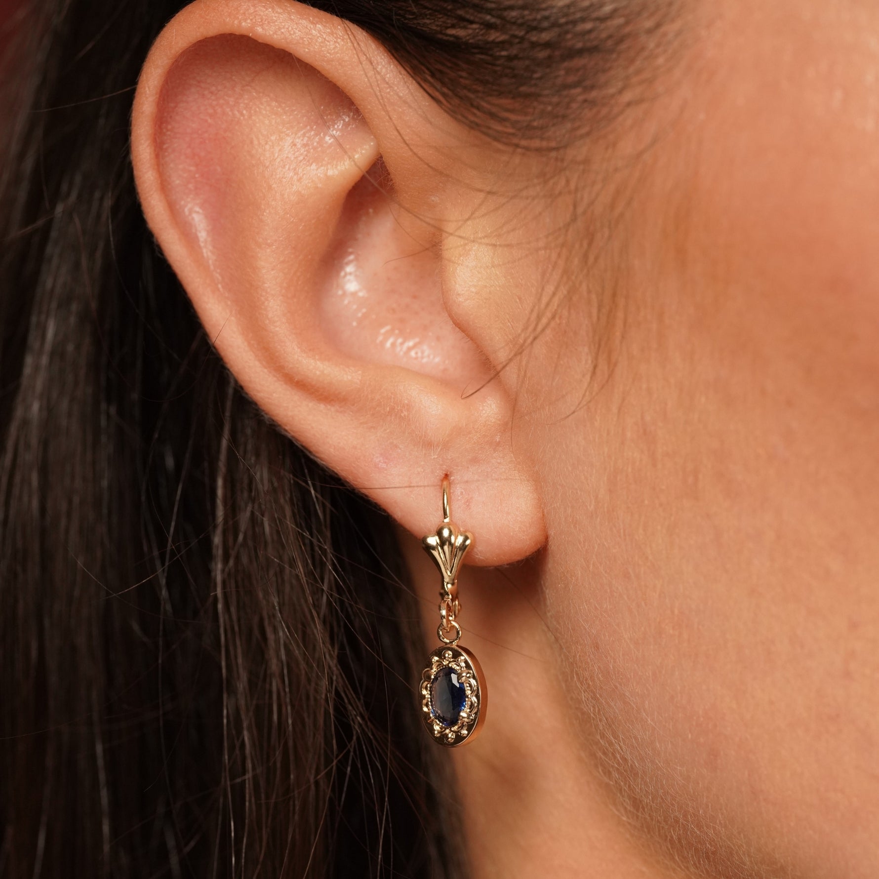 A close-up of a person's ear wearing a gold drop earring with an oval dark blue gemstone.