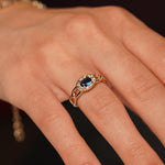 A close-up of a hand wearing a gold ring with a dark blue oval center stone and small clear accent stones around it.