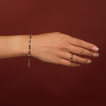 A hand wearing a delicate gold chain bracelet with small dark blue stones and a matching gold ring with a blue teardrop-shaped gemstone, set against a solid dark red background.