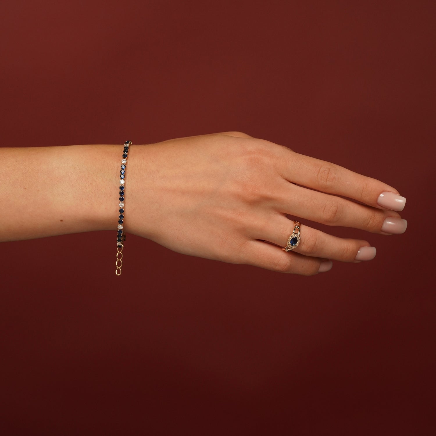 A hand wearing a delicate gold chain bracelet with small dark blue stones and a matching gold ring with a blue teardrop-shaped gemstone, set against a solid dark red background.