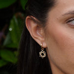 A close-up of a woman's ear wearing a small gold dangle earring with a flower-like openwork design.