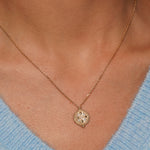 A close-up of a person's neck and chest wearing a delicate gold chain necklace with a round medallion pendant, paired with a light blue knit top.