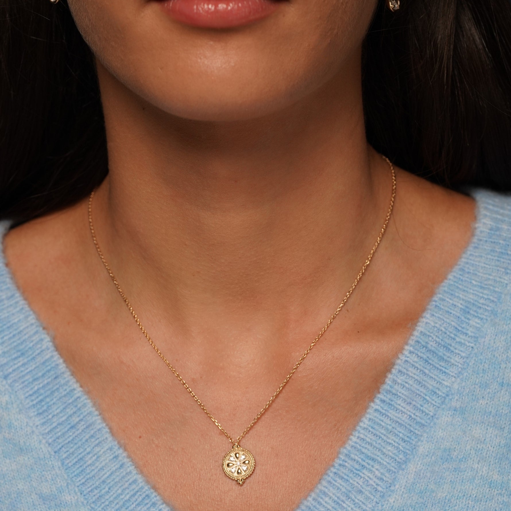 A close-up of a model wearing a delicate gold chain necklace with a small circular pendant featuring a floral or sunburst design with tiny clear stones, paired with a light blue knit sweater.