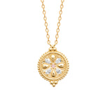 A gold pendant necklace featuring a circular, textured medallion with a floral starburst design and a small pink gemstone at the center, shown on a plain white background.
