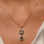 A close-up of a delicate gold chain necklace worn on a person's neck, featuring a double-drop pendant with a clear oval stone above a teardrop-shaped dark brown gemstone surrounded by small clear stones.