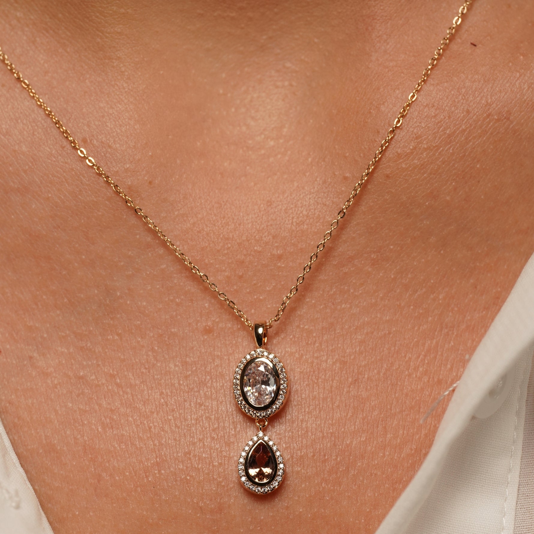 A close-up of a delicate gold chain necklace worn on a person's neck, featuring a double-drop pendant with a clear oval stone above a teardrop-shaped dark brown gemstone surrounded by small clear stones.