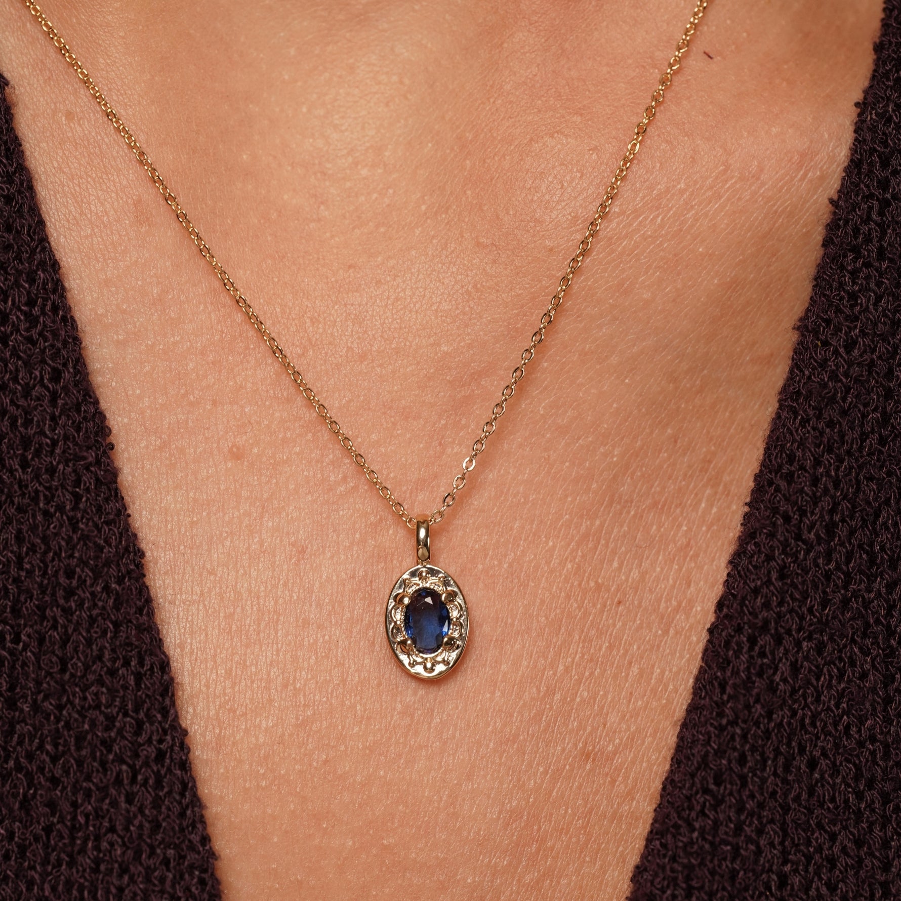 A close-up of a gold chain necklace with an oval pendant featuring a deep blue gemstone surrounded by a textured gold border, worn on a model's neck.