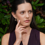A woman with dark hair wearing a delicate gold necklace with a small flower-like pendant, matching drop earrings, and a ring, posing in front of a green leafy background.