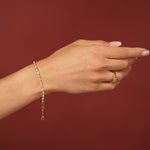 A delicate gold tennis bracelet with small clear stones worn on a woman's wrist, shown against a solid dark red background. The hand is also wearing a gold ring.