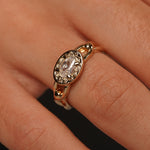 A close-up of a gold ring with an oval milky white gemstone and decorative filigree detailing, shown worn on a finger.