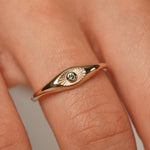 A close-up of a gold ring with an eye motif and a small central clear gemstone, worn on a person's finger.