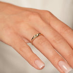 A close-up of a hand wearing a delicate gold ring with a small central gemstone on the ring finger, set against a soft, light beige background.