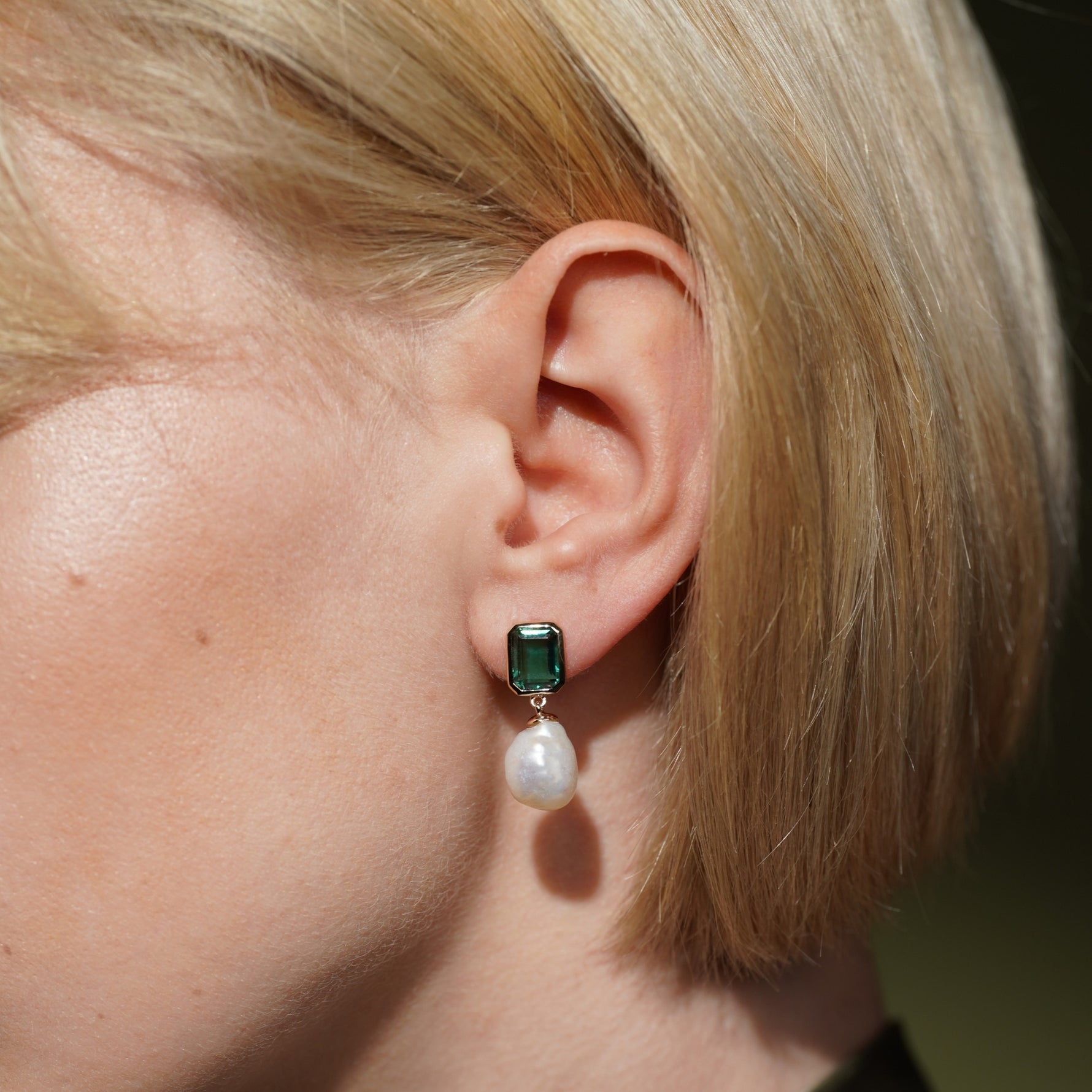 A close-up of a person's ear wearing a drop earring with a rectangular green gemstone and a dangling white pearl.